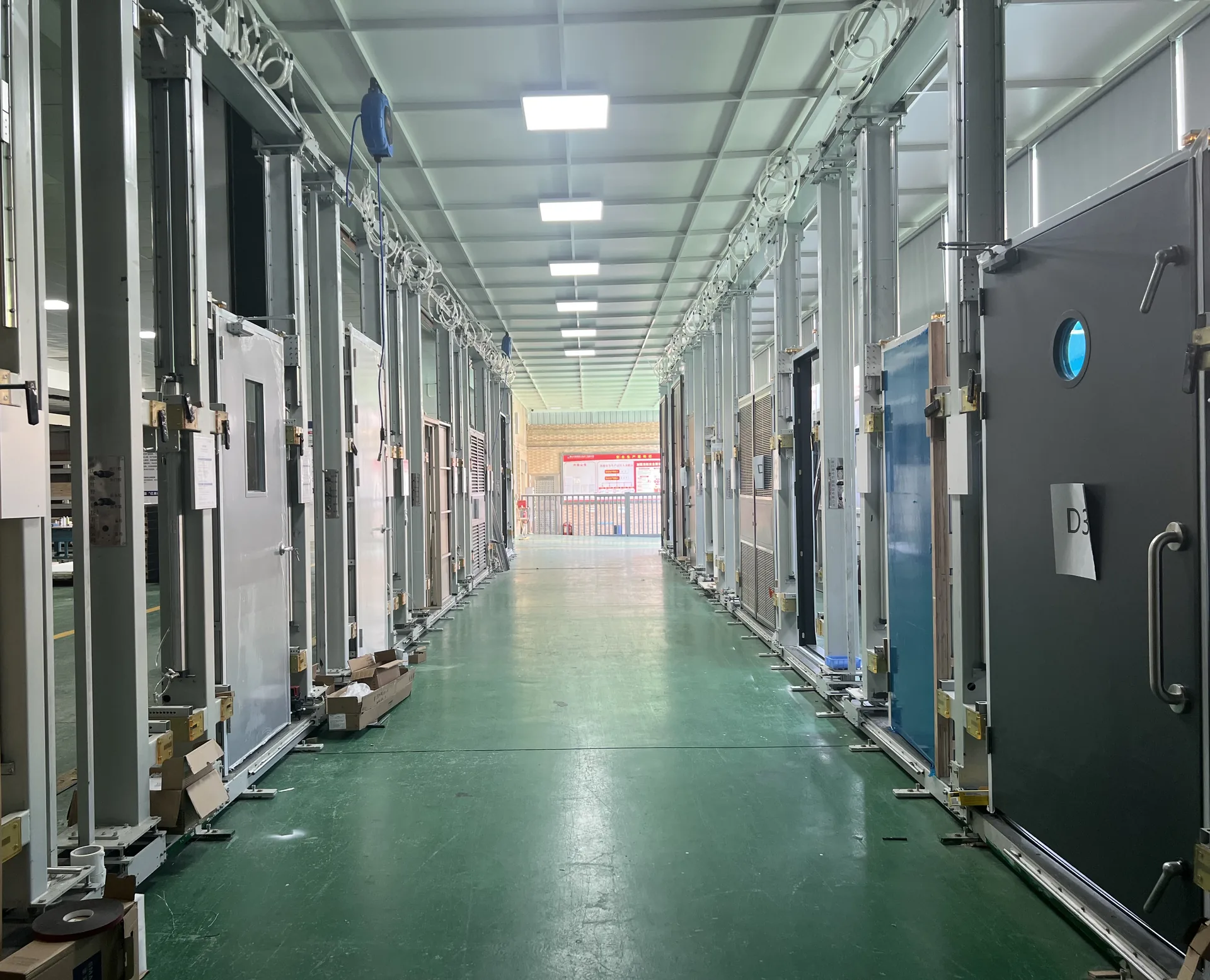 Door testing line in a factory: rows of steel and timber door leaves mounted in opening-and-closing test frames along a green-floored aisle