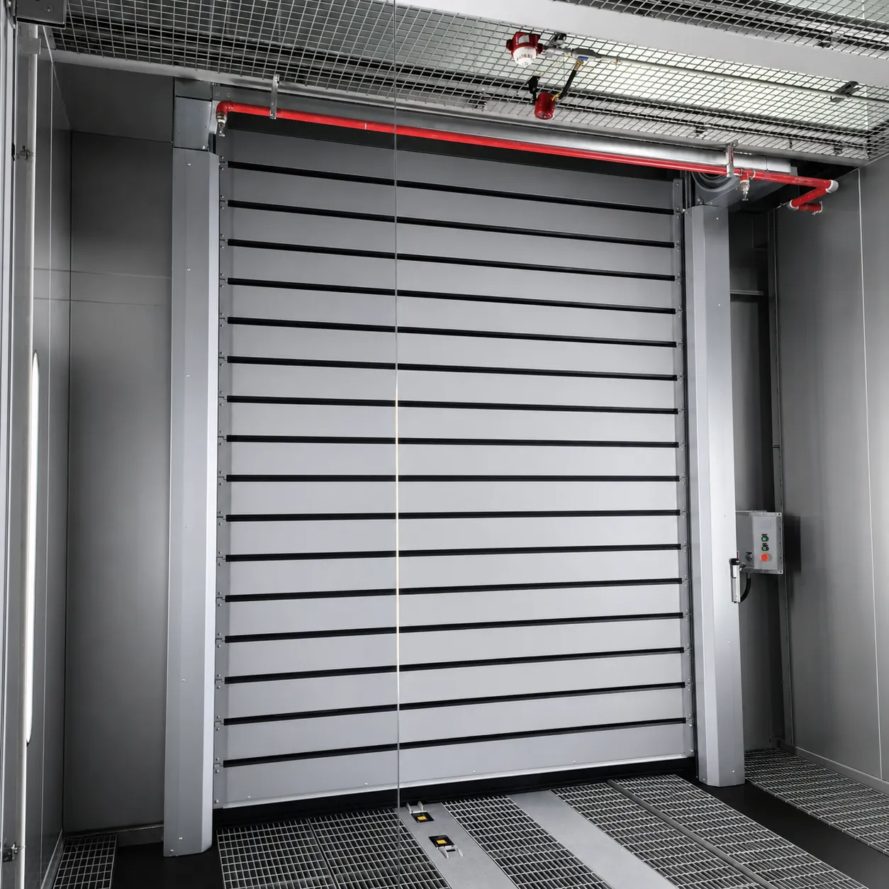 Light grey high speed spiral door at a booth entrance with grating floor, a red pipe above and a control box beside it