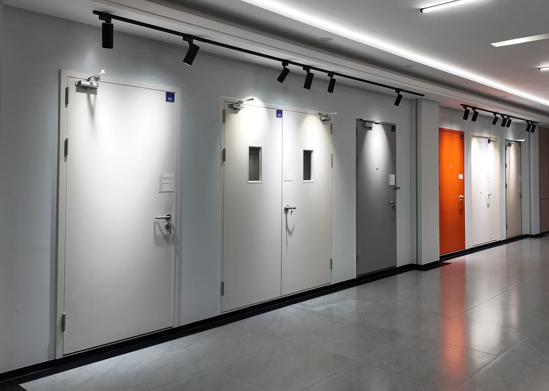 Showroom wall of steel doors: a white single door with closer, a white double door with vision panels, a grey single door, and orange, white leaf-and-a-half and beige doors further along