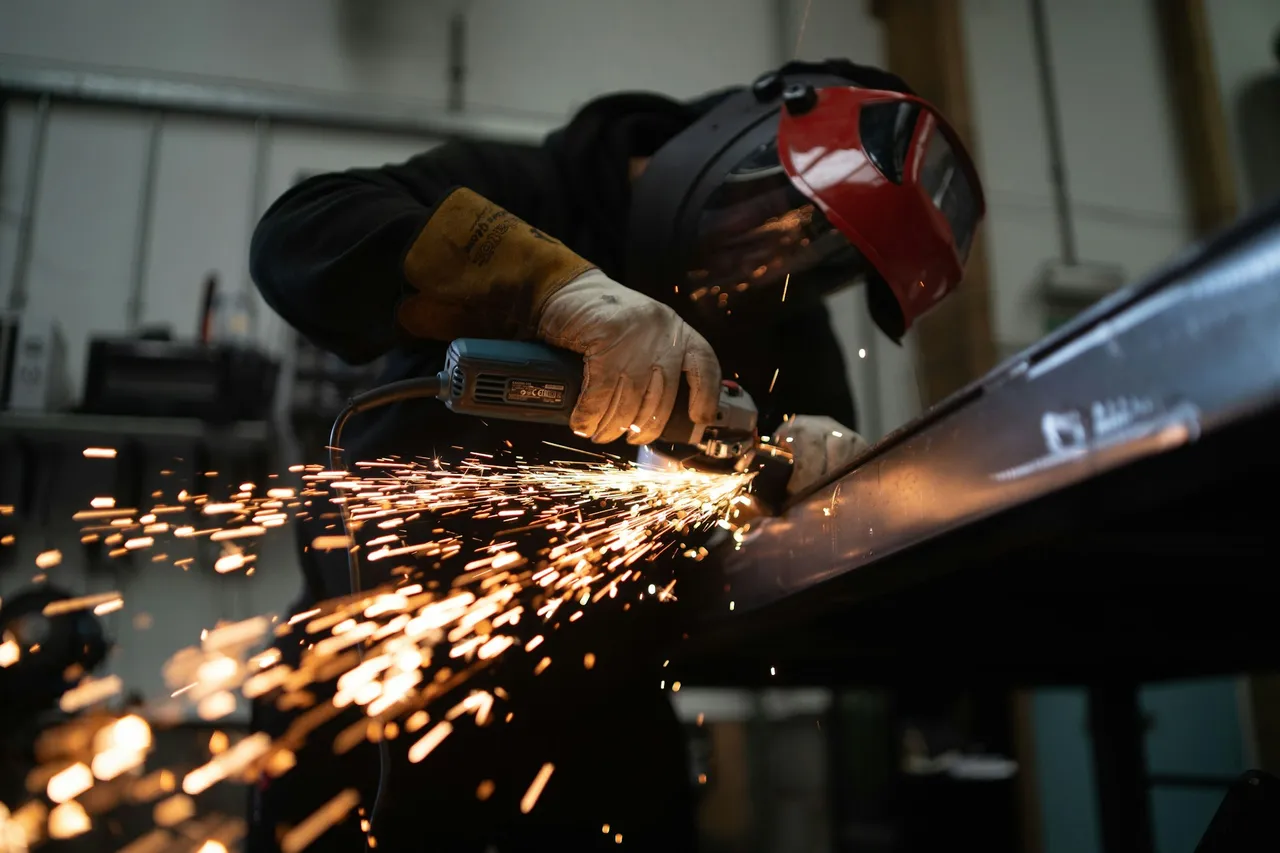 Worker in a welding helmet grinding a steel beam, with sparks flying